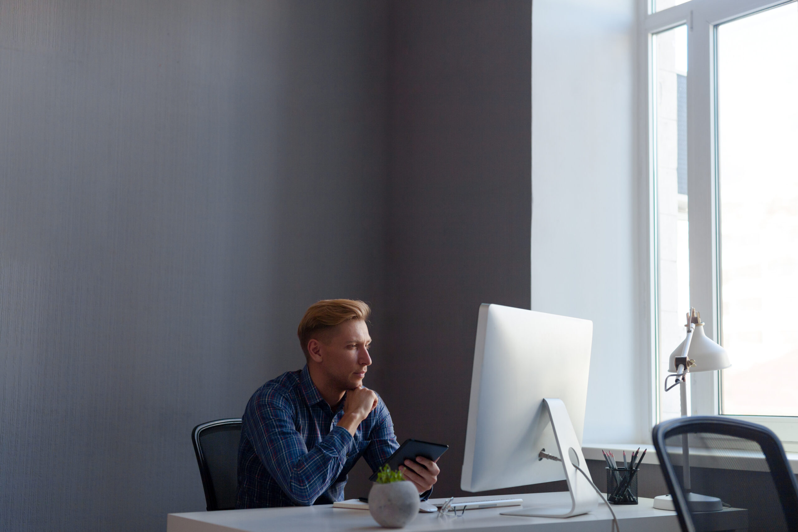 Young casual male sitting in front of computer with tablet in hands in office.